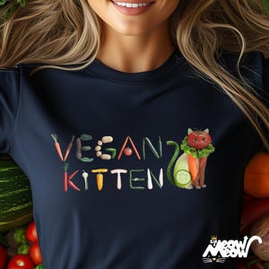 Puede incluir: Una camiseta azul marino con las palabras "VEGAN KITTEN" escritas con diferentes verduras. Un gato hecho de verduras lleva un collar de lechuga y sostiene una zanahoria.