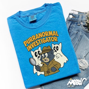 May include: A light blue t-shirt with the words "PURRANORMAL INVESTIGATOR" in yellow. The graphic features a cartoon cat detective with two ghost companions. The shirt is folded next to a pair of blue jeans.