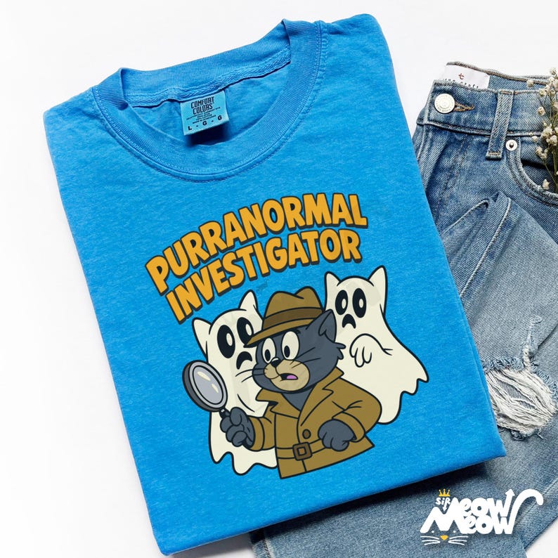 May include: A light blue t-shirt with the words "PURRANORMAL INVESTIGATOR" in yellow. The graphic features a cartoon cat detective with two ghost companions. The shirt is folded next to a pair of blue jeans.