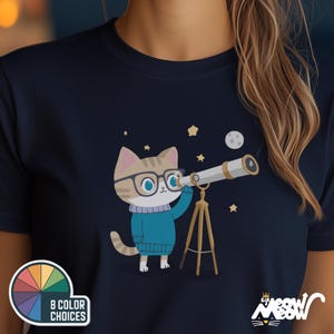 May include: A navy blue t-shirt featuring a cartoon illustration of a cat wearing glasses and looking through a telescope. The cat is surrounded by stars and a crescent moon. The text "8 Color Choices" is visible in the bottom left corner of the image.