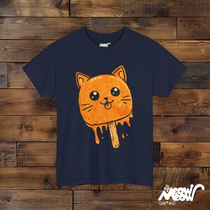 May include: A navy blue t-shirt with a cartoon orange cat popsicle graphic. The cat has a tongue sticking out and dripping orange ice cream.