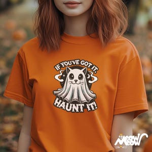 以下が含まれることがあります： オレンジ色のTシャツ。漫画のゴーストキャットのデザインが特徴です。デザインには、遊び心のあるフォントで「IF YOU'VE GOT IT. HAUNT IT！」というテキストが含まれています。このシャツは、ハロウィーンにぴったりの無地のオレンジ色です。
