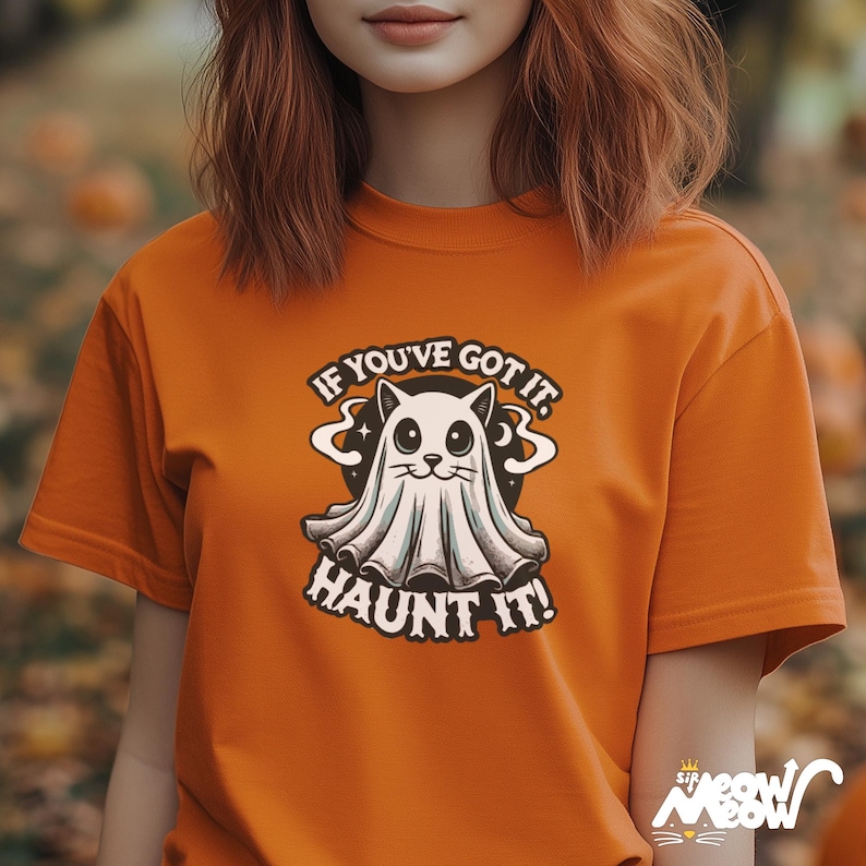 以下が含まれることがあります： オレンジ色のTシャツ。漫画のゴーストキャットのデザインが特徴です。デザインには、遊び心のあるフォントで「IF YOU'VE GOT IT. HAUNT IT！」というテキストが含まれています。このシャツは、ハロウィーンにぴったりの無地のオレンジ色です。