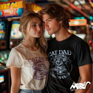 Cat Parent Gamer Club T-Shirts. Photo shown is a man and woman. The woman is wearing the ivory "Cat Mom" shirt whilst the man is wearing the black "Cat Dad" tee. Both tees show a cat holding a gaming controller.