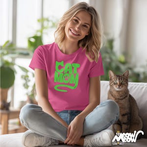 May include: A magenta t-shirt with the text "CAT MOM" in green, featuring a paw print. A tabby cat sits on a white couch next to the person wearing the shirt. The shirt is a crew neck style.