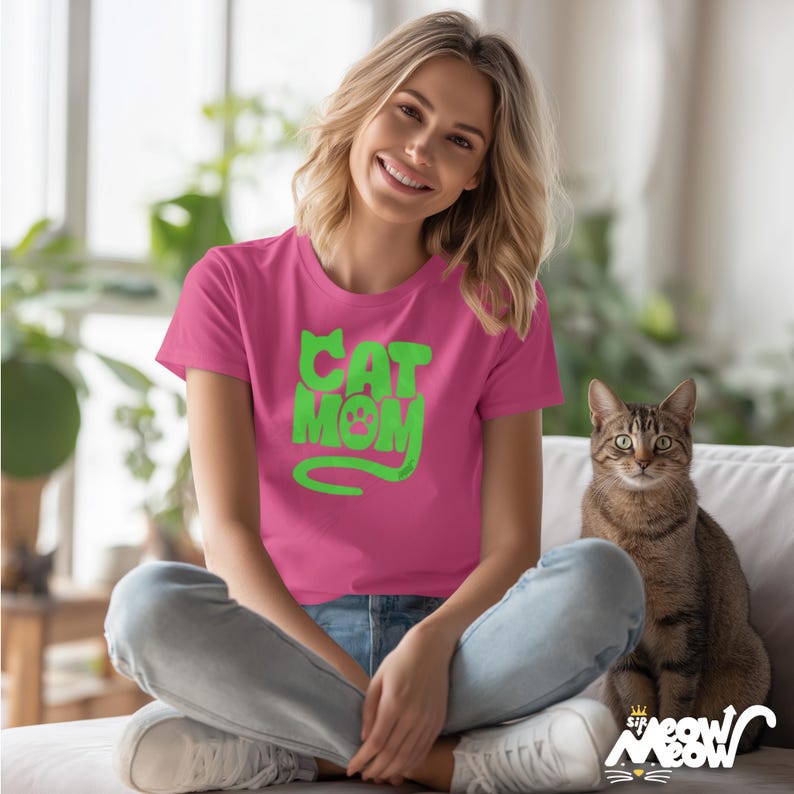 May include: A magenta t-shirt with the text "CAT MOM" in green, featuring a paw print. A tabby cat sits on a white couch next to the person wearing the shirt. The shirt is a crew neck style.