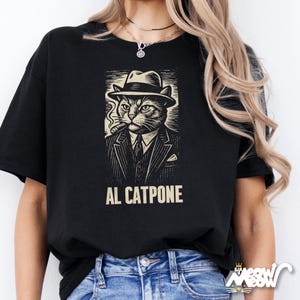 K&ouml;nnte beinhalten: Schwarzes T-Shirt mit einer detaillierten Illustration einer Katze mit Hut und Anzug, die eine Zigarre raucht. Der Text "AL CATPONE" ist unter dem Bild aufgedruckt. Das Shirt ist ein bequemer, l&auml;ssiger Stil.