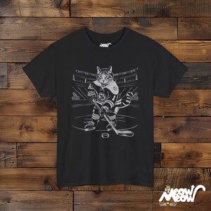 Op de afbeelding: Zwart T-shirt met een afbeelding van een kat die hockey speelt. De kat draagt een hockeytrui en houdt een hockeystick vast. De tekst "Sir Meow" staat gedrukt in de rechterbenedenhoek van het shirt.