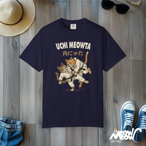 K&ouml;nnte beinhalten: Ein dunkelblaues Kurzarm-T-Shirt mit einer Grafik von zwei Katzen in wei&szlig;en Kampfkunst-Uniformen mit schwarzen G&uuml;rteln. &Uuml;ber den Katzen sind der Text UCHI MEOWTA sowie japanische Schriftzeichen aufgedruckt, die einen Wurf darstellen.