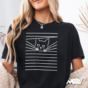 May include: Black t-shirt with a white graphic of a cat peeking through blinds. The cat has a sad expression and is looking directly at the viewer.