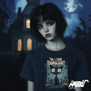 May include: A woman with black hair and dark makeup is wearing a dark blue t-shirt with a black cat graphic and the text "Do I Look Familiar?" The cat is in a graveyard setting with tombstones and a pumpkin.