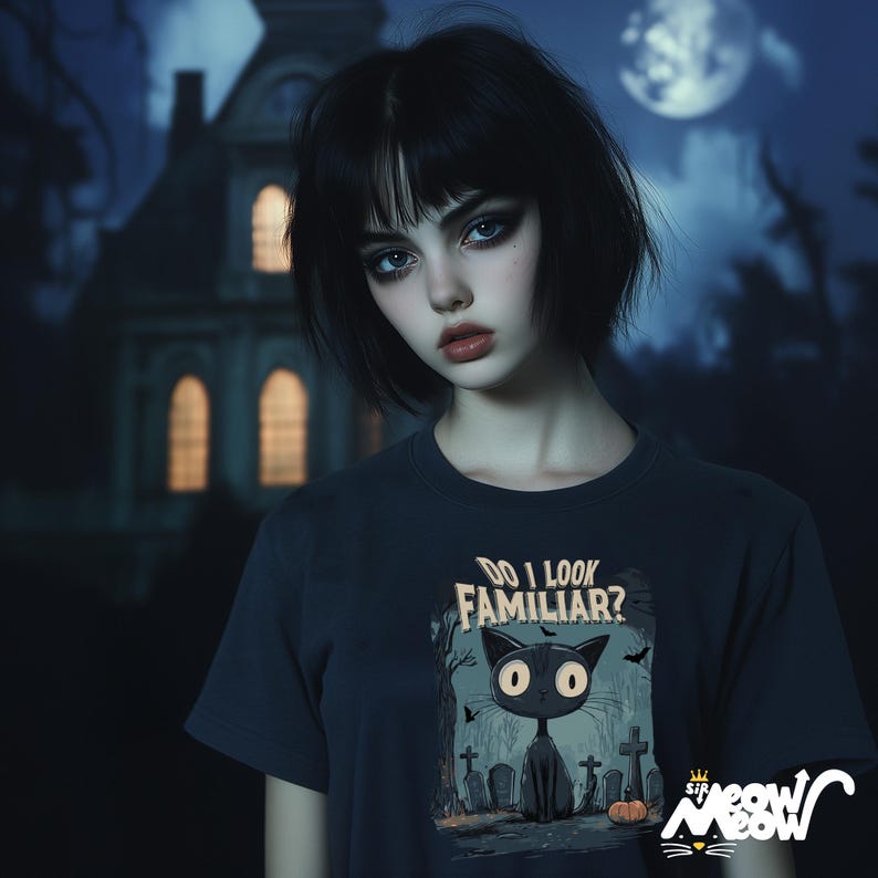May include: A woman with black hair and dark makeup is wearing a dark blue t-shirt with a black cat graphic and the text "Do I Look Familiar?" The cat is in a graveyard setting with tombstones and a pumpkin.