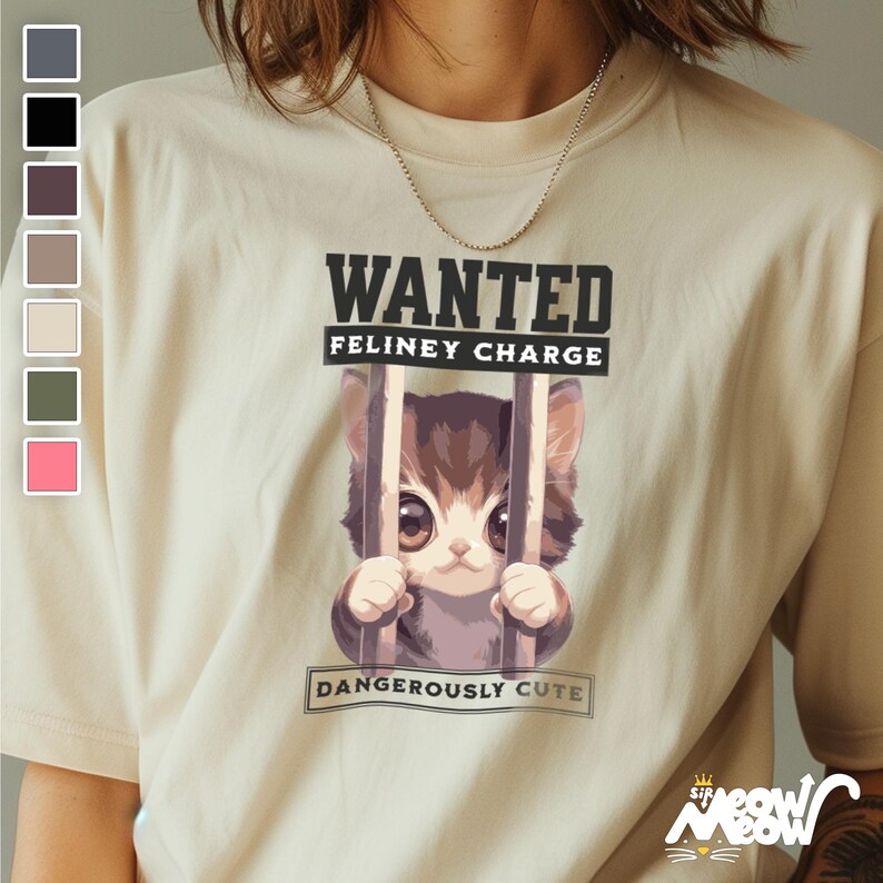 May include: A beige t-shirt with a graphic of a cartoon cat in a cage. The text "WANTED FELINEY CHARGE DANGEROUSLY CUTE" is printed on the shirt.