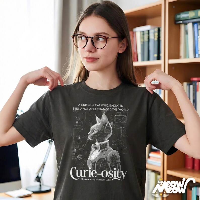 May include: Dark gray t-shirt featuring a detailed illustration of a cat in a suit and bow tie, with the text "Curie-osity" and "A Curious Cat Who Radiated Brilliance and Changed the World". The shirt is a crew neck.