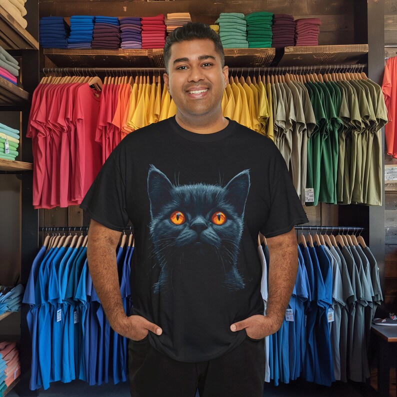 May include: Black t-shirt featuring a detailed illustration of a black cat with bright orange eyes. The cat's fur is depicted with shading, giving it a realistic appearance. The t-shirt is displayed in a retail setting with racks of colorful t-shirts in the background.