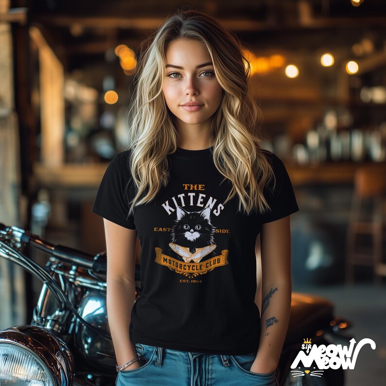 K&ouml;nnte beinhalten: Schwarzes T-Shirt mit einem Katzen-Motiv und der Aufschrift "The Kittens Motorcycle Club". Das Design beinhaltet ein Banner mit dem Text "East Side" und "Est. 1984". Das T-Shirt wird mit einer blauen Jeans getragen.