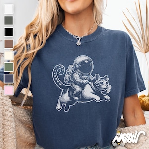 Denim colored Comfort Colors 1717 unisex T-shirt with a humorous white graphic of an astronaut riding atop a playful cat, combining outer space and cat lover themes.