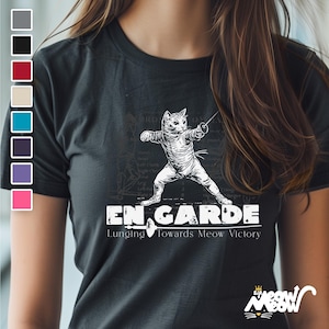 Może przedstawiać: Czarny T-shirt z białym nadrukiem przedstawiającym kota szermierza. Pod kotem znajduje się napis "En Garde", a pod nim napis "Lunging Towards Meow Victory".
