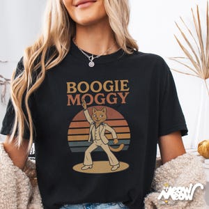 May include: Black t-shirt with the text "Boogie Moggy" above a cartoon cat in a white suit, striking a disco pose against a sunset backdrop. The cat is on a dance floor. The shirt has a relaxed fit.