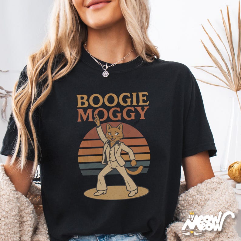 May include: Black t-shirt with the text "Boogie Moggy" above a cartoon cat in a white suit, striking a disco pose against a sunset backdrop. The cat is on a dance floor. The shirt has a relaxed fit.