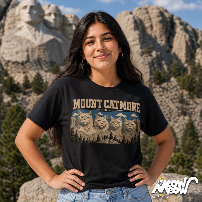 May include: Black t-shirt with the text "MOUNT CATMORE" above a graphic of four cat faces carved into a mountain, similar to Mount Rushmore. The shirt also has the text "sip meow meow" in the bottom right corner.