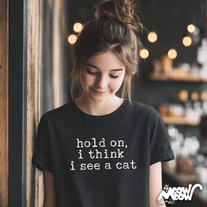 May include: Dark gray t-shirt with the text "hold on, i think i see a cat" in white. The shirt has short sleeves and a crew neck. The bottom right corner has a logo with a cat and the words "Sir Meow".
