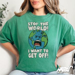 Könnte beinhalten: Grünes T-Shirt mit dem Text "STOP THE WORLD! I WANT TO GET OFF!" und einer Darstellung der Erde mit einem schreienden Gesicht. Die Grafik zeigt blaue Ozeane und grüne Kontinente. Das Shirt hat kurze Ärmel und einen Rundhalsausschnitt.