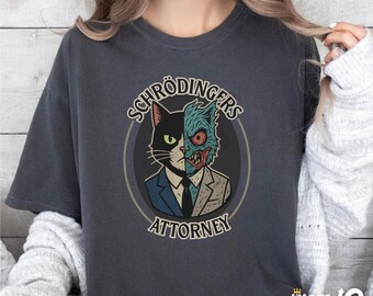 May include: A dark gray t-shirt with a graphic of a cat with a split face. The cat's left side is black and the right side is a green monster. The text "Schrödinger's Attorney" is printed below the cat.