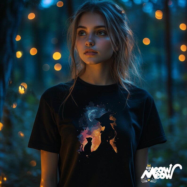 May include: A woman with long brown hair is wearing a black t-shirt featuring a silhouette of a witch and a cat in front of a moon. The design is in shades of orange, yellow, and blue.