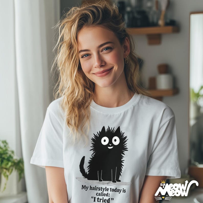 May include: White t-shirt featuring a cartoon black cat with the text "My hairstyle today is called: I tried". The cat has large white eyes and a smiling expression. The shirt is worn by a person with blonde hair.