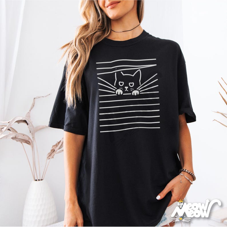 May include: Black oversized t-shirt with a white graphic of a cat peeking through horizontal lines. The cat has a simple design with a round head, two ears, and a small mouth.
