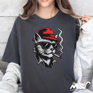 May include: Dark grey t-shirt with a cat illustration. The cat is depicted wearing a red fedora, sunglasses, and smoking a cigarette. The design is in black, white, and red. The shirt has the text "Sip Meow" at the bottom.