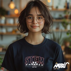 May include: A woman with short brown hair wearing round glasses and a black t-shirt with the text "STEM Science Teachers Encourage MEOWS" in red and white. The shirt also has a paw print and cat silhouettes.