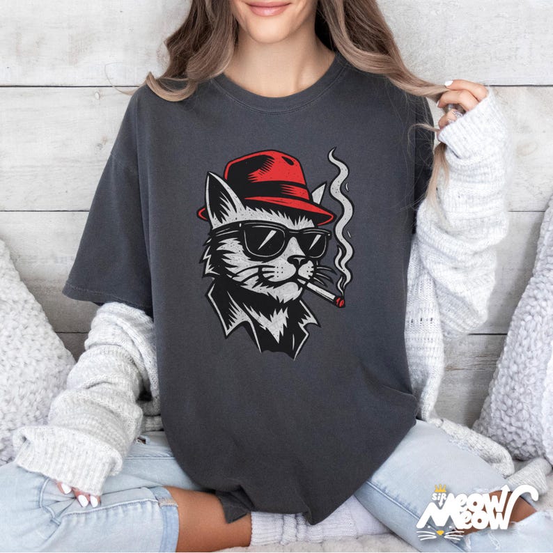 May include: Dark grey t-shirt with a graphic of a cat in a red hat and sunglasses, smoking a cigarette. The cat is in black and white. The text "Sip Meow" is at the bottom of the shirt.