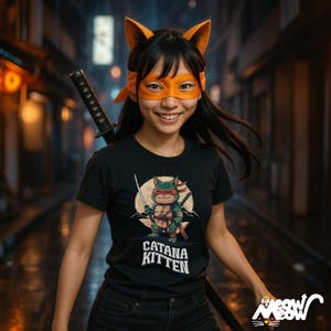 May include: A person wearing a black t-shirt with a cartoon cat graphic holding a sword, with the text "CATANA KITTEN". The person is also wearing an orange mask and headband with cat ears, and a katana sword is visible.