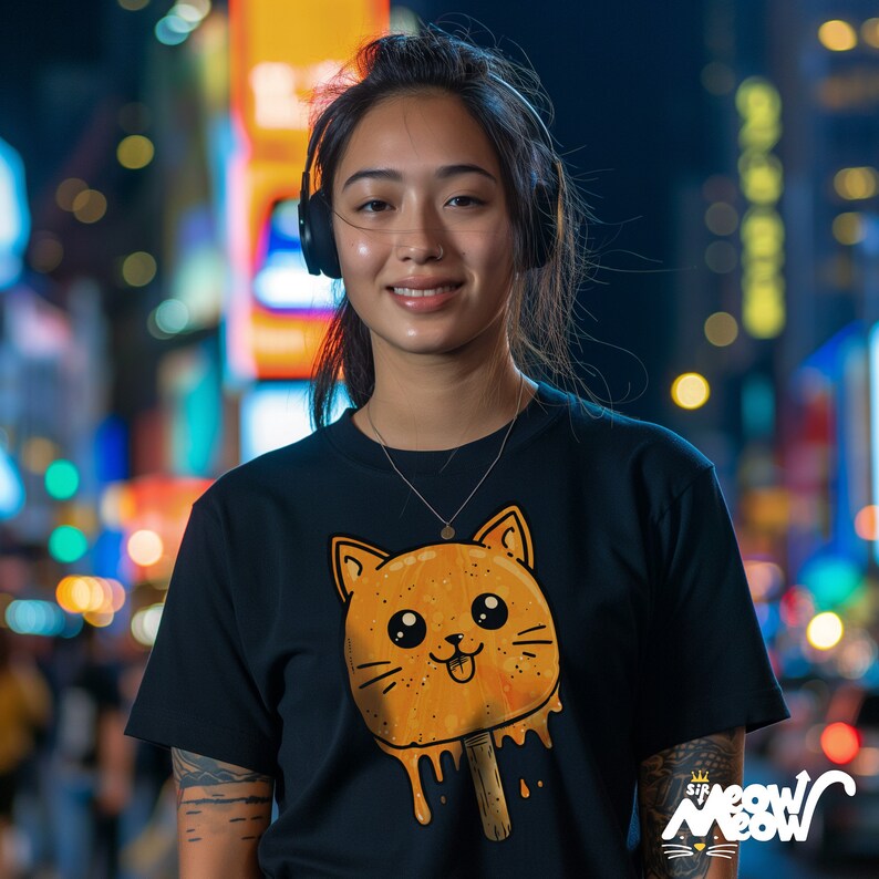 May include: A woman wearing a black t-shirt with a cartoon orange cat popsicle graphic. The graphic has a dripping effect and the text "Sir Meow Meow" is visible in the bottom right corner of the image.