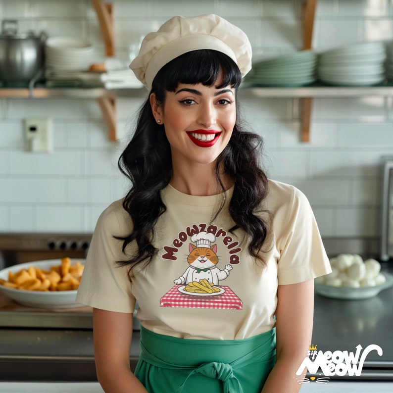 Puede incluir: Una mujer con un gorro de cocinero blanco y una camiseta crema con un gato de dibujos animados con un gorro de cocinero y sosteniendo un plato de patatas fritas. El texto de la camiseta dice "Meowzarella".