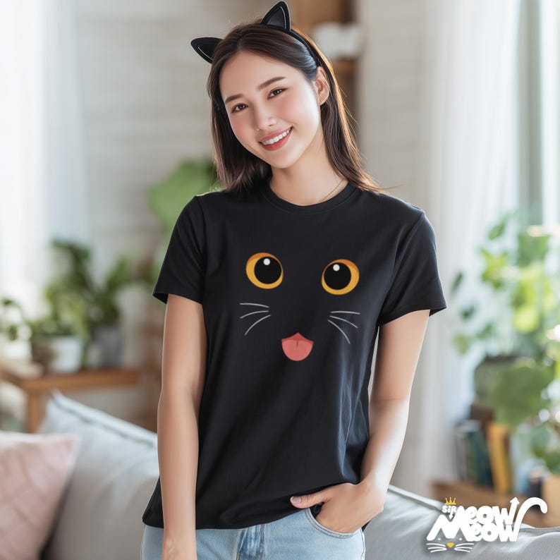 May include: Black t-shirt featuring a cat face design with large yellow eyes, whiskers, and a pink tongue. The shirt has a round neckline and short sleeves. The image also includes a black cat ear headband.