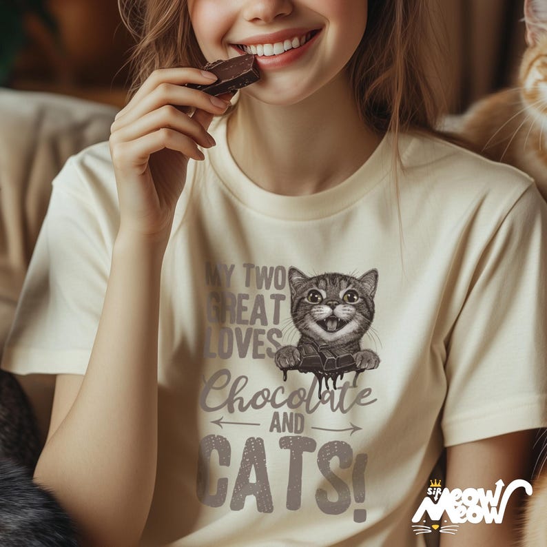 May include: A woman wearing a cream colored t-shirt with a graphic of a cat eating chocolate and the text "My two great loves Chocolate and Cats!"