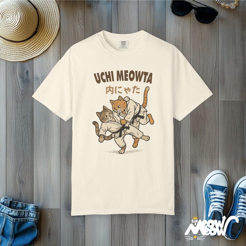 K&ouml;nnte beinhalten: Ein cremefarbenes Kurzarm-T-Shirt mit einer Grafik von zwei Katzen in wei&szlig;en Kampfkunst-Uniformen mit schwarzen G&uuml;rteln. Der Text UCHI MEOWTA und japanische Schriftzeichen sind in dunkelbrauner Schrift &uuml;ber den Katzen gedruckt.