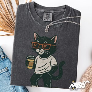 May include: Dark grey Comfort Colors t-shirt with a cartoon cat wearing sunglasses and holding a coffee cup. The cat is dressed in a white t-shirt and dark trousers. A silver necklace and dried foliage are also visible.