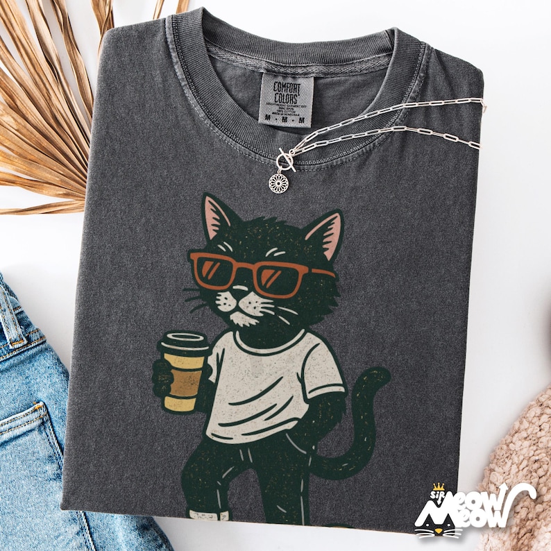 May include: Dark grey Comfort Colors t-shirt with a cartoon cat wearing sunglasses and holding a coffee cup. The cat is dressed in a white t-shirt and dark trousers. A silver necklace and dried foliage are also visible.