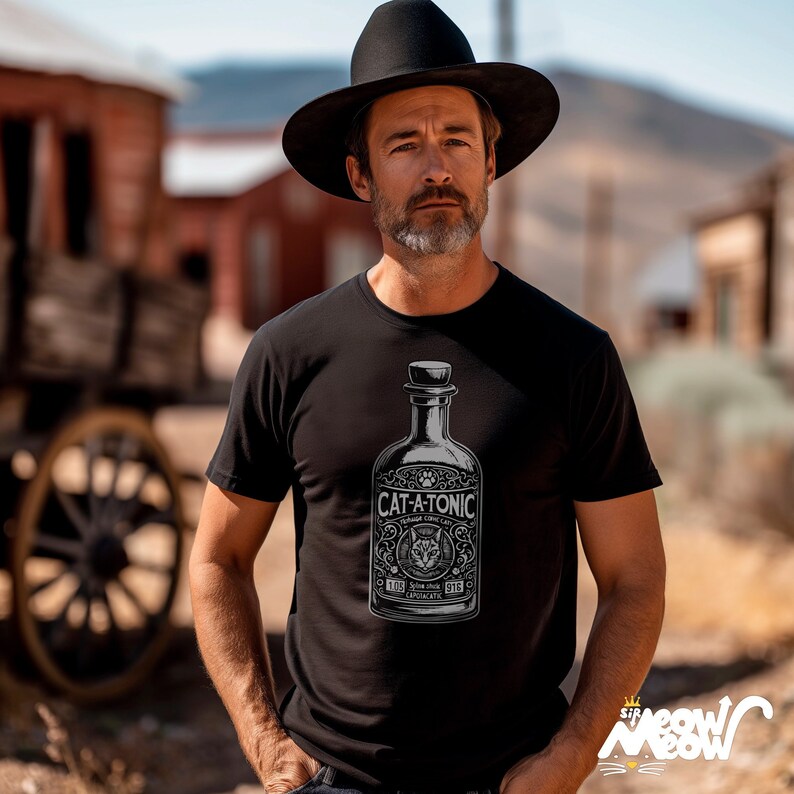 May include: Black t-shirt featuring a vintage-style graphic of a bottle labeled "CAT-A-TONIC". The design includes a cat illustration, paw prints, and decorative elements. The shirt is worn by a person wearing a black hat.