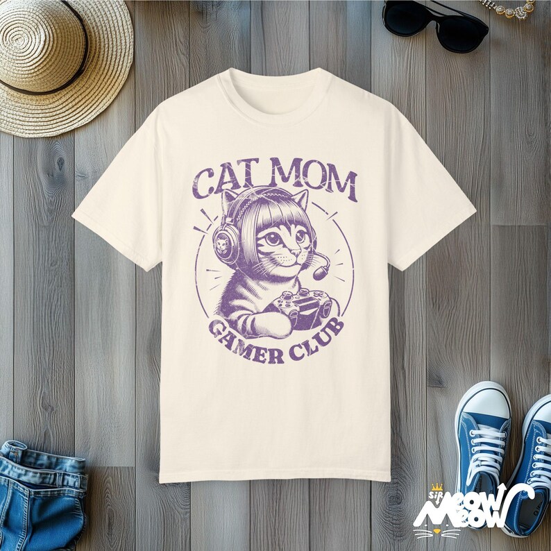 Cat Parent Gamer Club T-Shirts. The tee shows a cat holding a gaming controller. Above the image is the text "Cat Mom" and below the image is the text "Gamer Club".
The t-shirt shown is in the color "Ivory" and is also available in a "Cat Dad" version.