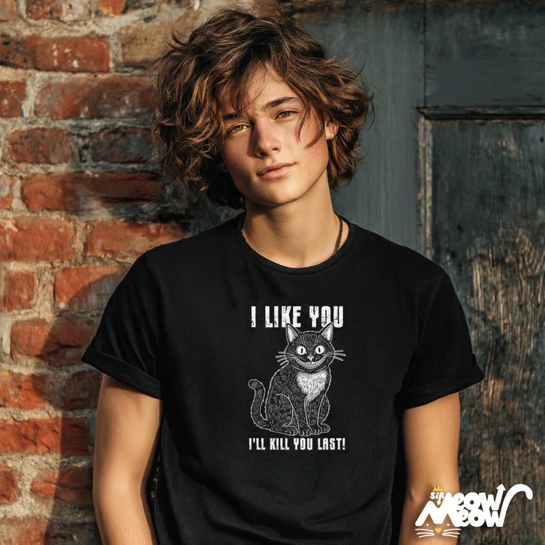 May include: Black t-shirt with the text "I LIKE YOU" above a cartoon cat illustration and the text "I'LL KILL YOU LAST!". The cat has a white chest and a sinister grin. The shirt is worn by a person.