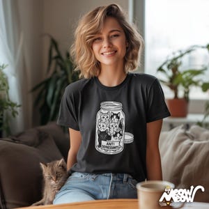 May include: Black t-shirt featuring a graphic of several cats inside a jar labeled "Anti Depressants." The design is in black and white. The shirt is worn by a person.