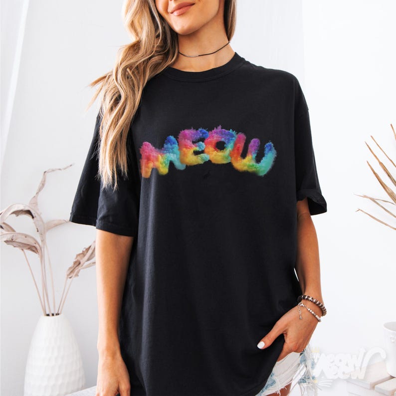 K&ouml;nnte beinhalten: Schwarzes T-Shirt mit dem Wort "MEOW" in einer bunten, regenbogenfarbenen, flauschigen Schrift. Das T-Shirt hat kurze &Auml;rmel und eine lockere Passform. Das Design befindet sich mittig auf der Vorderseite des Kleidungsst&uuml;cks.