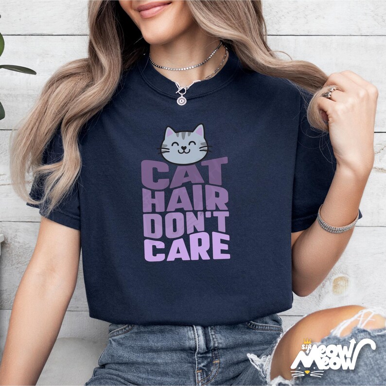 Pode incluir: Camiseta azul marinho com a frase "CAT HAIR DON'T CARE" em roxo e um desenho de gato cinza. A camiseta &eacute; de gola redonda.