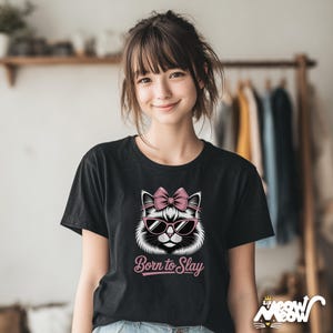 May include: Black t-shirt featuring a white cat graphic wearing pink sunglasses and a bow. The text "Born to Slay" is below the cat. The shirt is worn by a person.