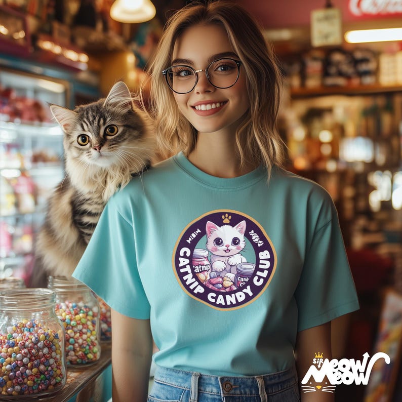 May include: A woman wearing a light blue t-shirt with a purple and gold catnip candy club logo featuring a cartoon cat. The logo says "Meow Catnip Candy Club". The woman is holding a white cat on her shoulder.